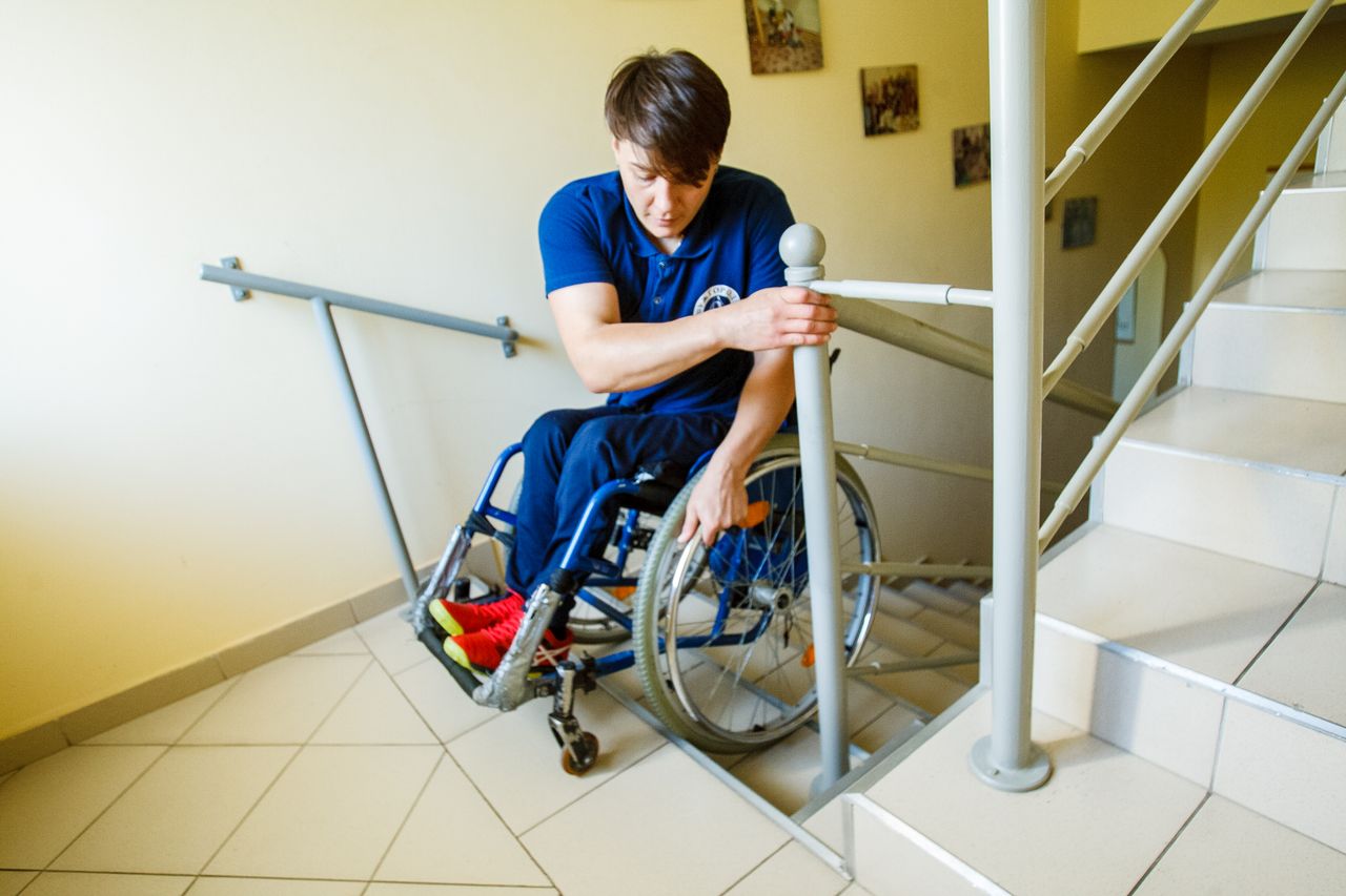 A man in a wheelchair using a staircase railing for support indoors.
