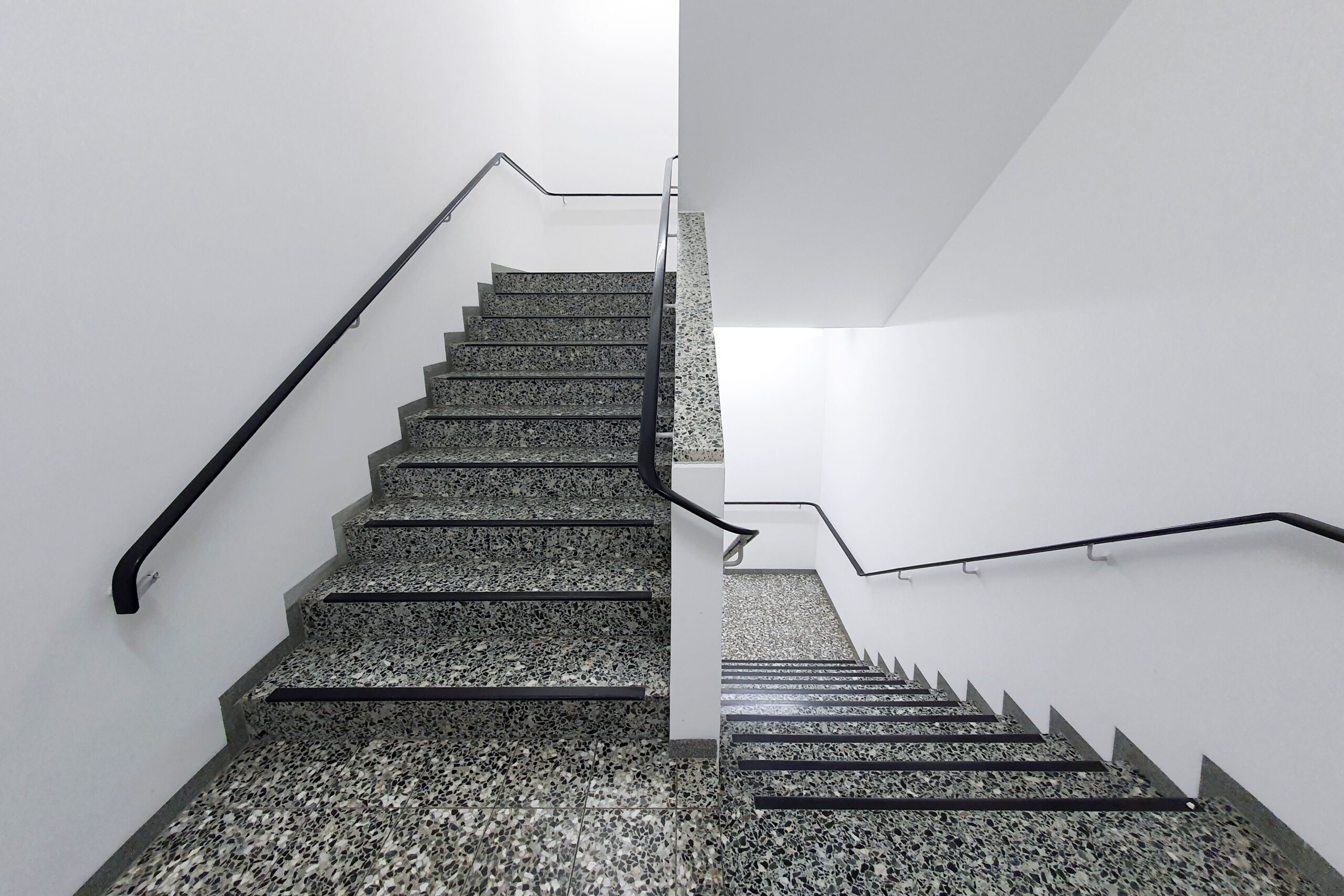 Treppenlift für gerade Treppen – Kosten, Vorteile, Einbau und wichtige Kaufkriterien Minimalist interior view of a granite staircase with black handrails, featuring clean white walls.