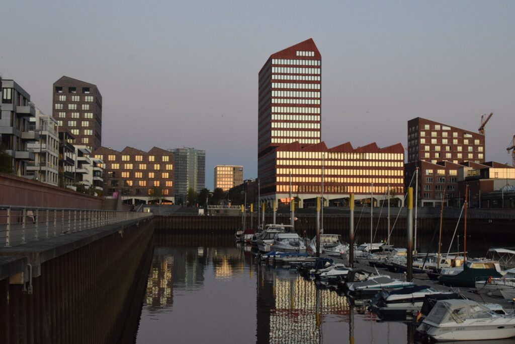 Skyline of modern buildings reflecting in the water at sunset, with boats in Bremen's harbor.