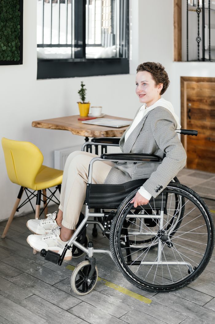 Side view full body of positive female freelancer in wheelchair sitting near table with laptop and coffee while spending time in cafeteria