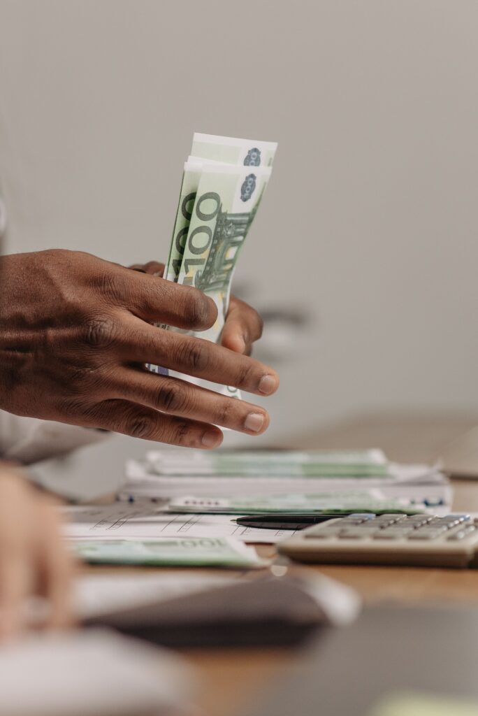 pexels photo 7654164 7654164 Close-up of hands counting US dollar bills on a cluttered desk in an office setting.