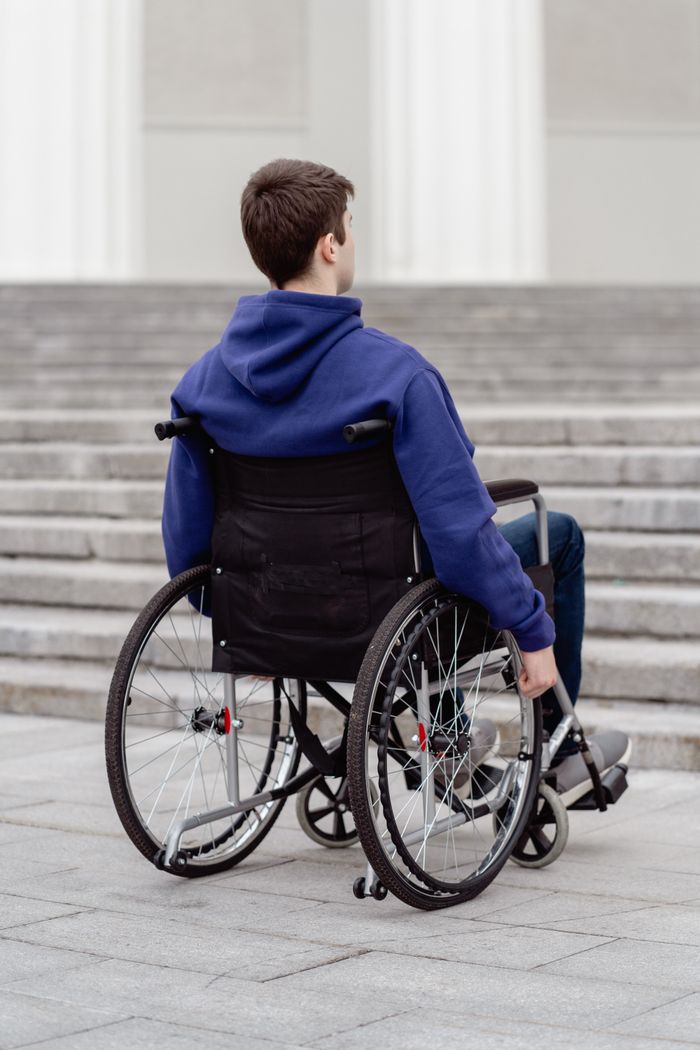 services-03 A young man in a wheelchair with a blue hoodie facing a staircase outdoors.