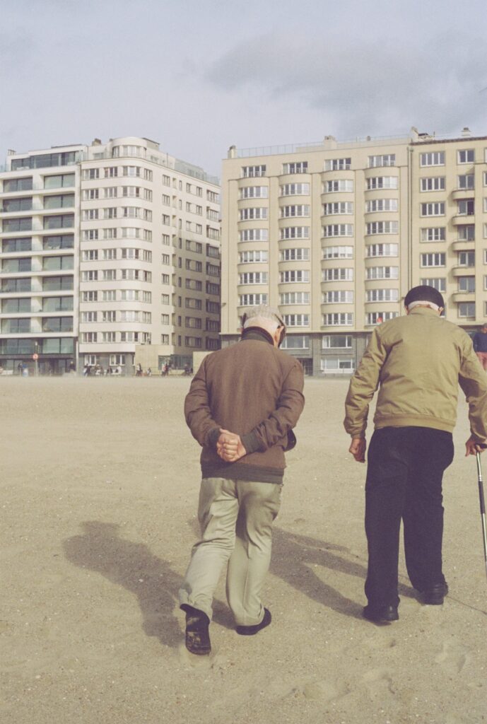 Treppenlift langfristig nutzen Bochum – Wartung und Lebensdauer Two senior men walking on a sandy urban beach with modern buildings in the background.