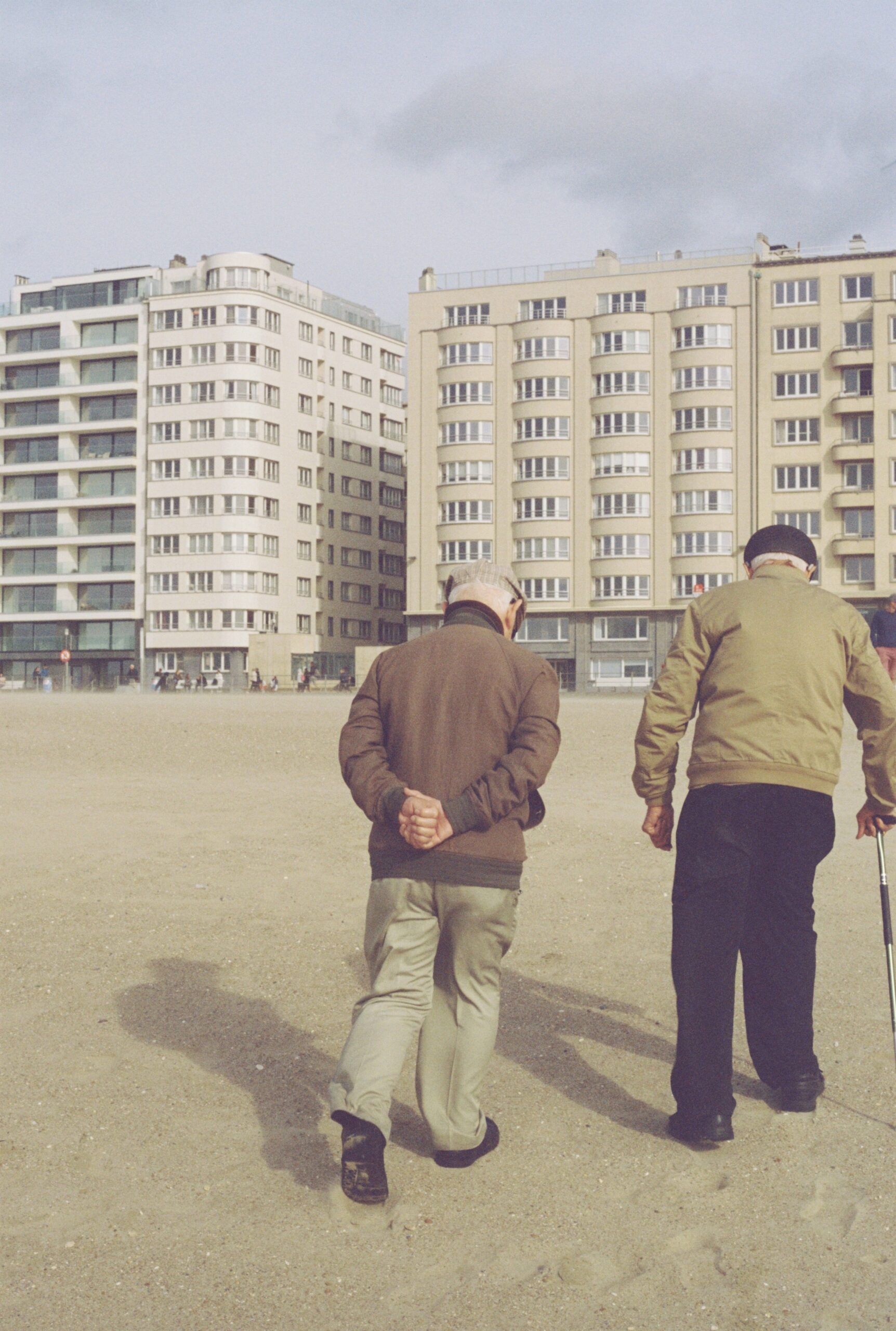 Treppenlift langfristig nutzen Bochum – Wartung und Lebensdauer Two senior men walking on a sandy urban beach with modern buildings in the background.